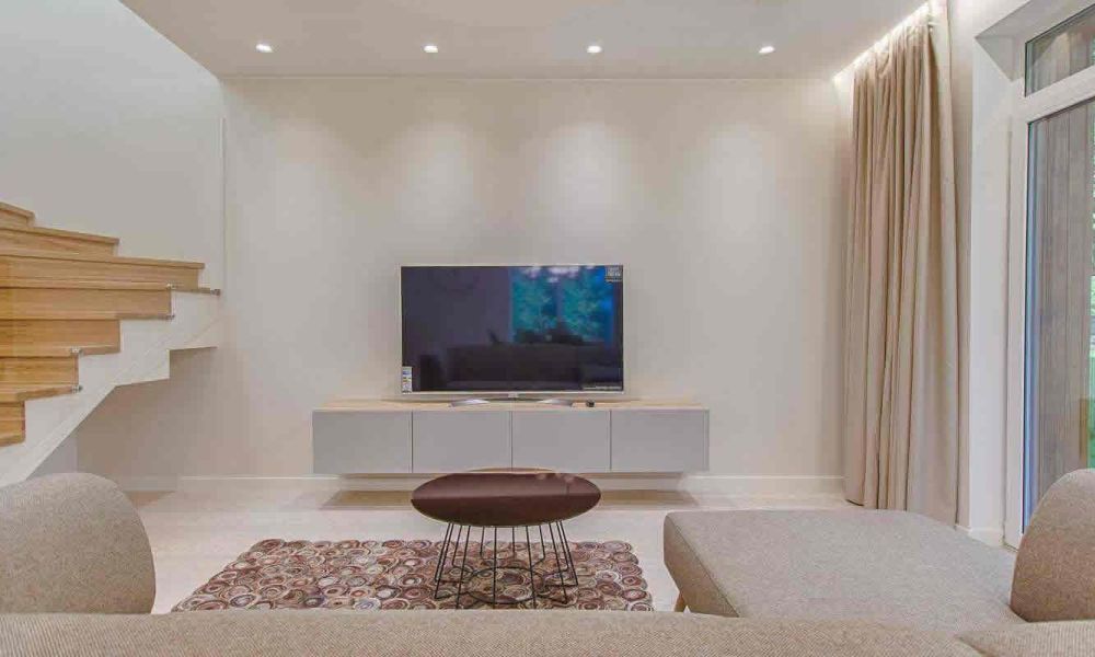 A modern living area with a beige sectional sofa, a flat-screen TV, a wooden staircase, and large windows, offering a bright and open design.