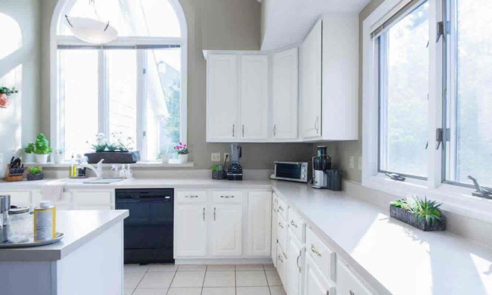 A bright kitchen with white cabinets, a large window above the sink, and various kitchen appliances on the counters, creating a clean and airy atmosphere.