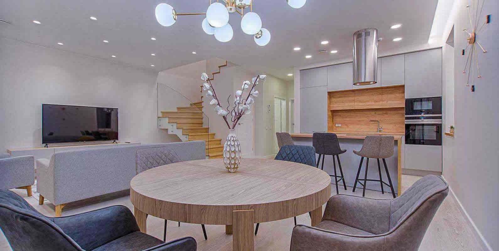 An open-concept living and dining area with a round dining table, gray chairs, a staircase, and a modern kitchen, showcasing a blend of functionality and style.